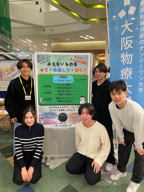 「みえないものをみて・体感して・知ろう」の案内ボードの前に立つボランティアサークルの学生5人